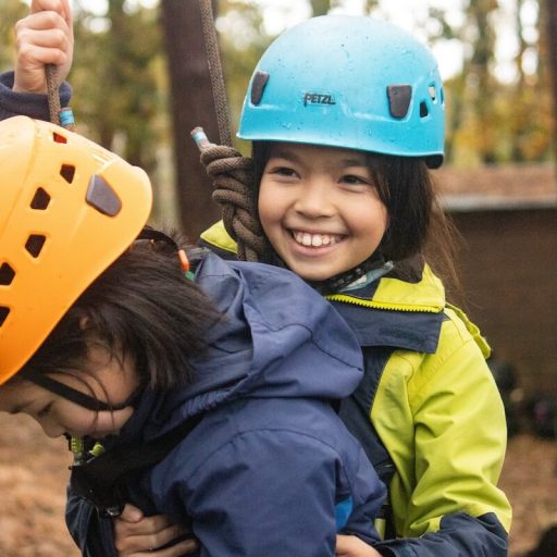 Two young people wearing helmets and coats attached to ropes with harness smiling
