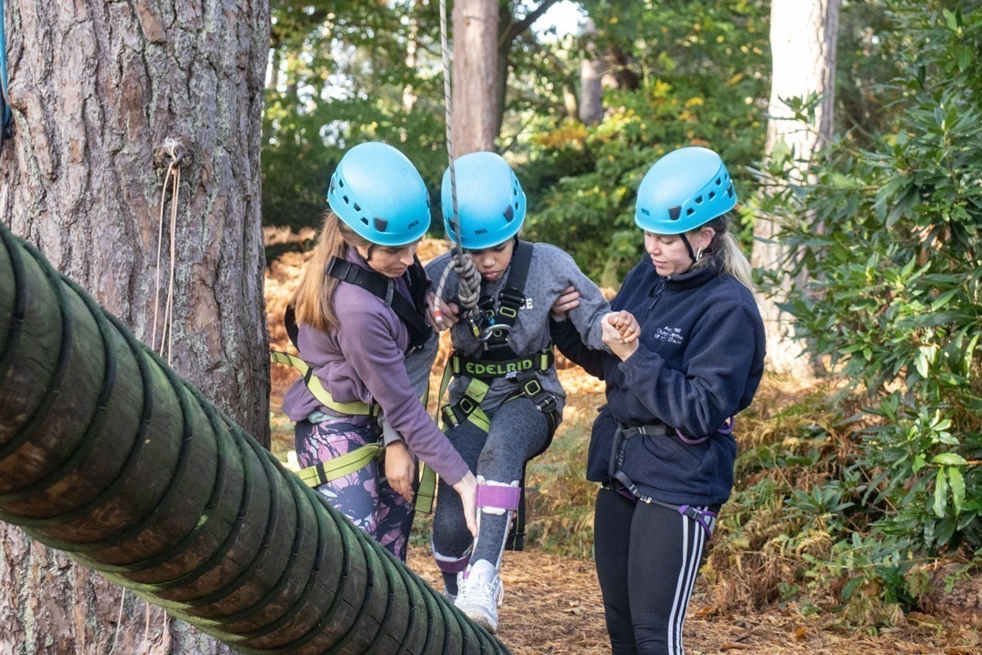 Young girl wearing blue helmet and harness strapped to rope walking up wooden beam holding on to adult and instructor doing high ropes all wearing helmets