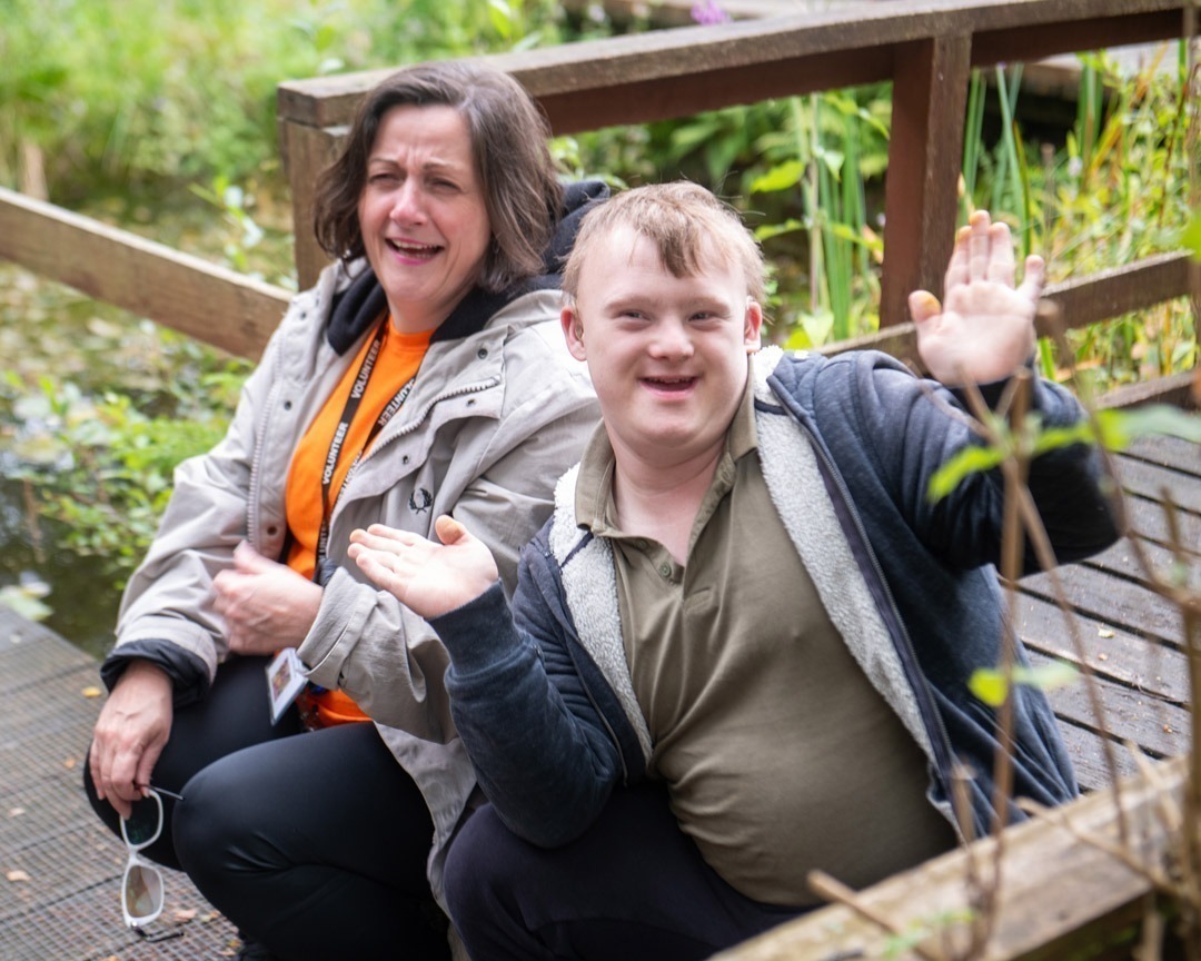 Man with lady sat on a wooden decking with plants all around them smiling to camera - man posing
