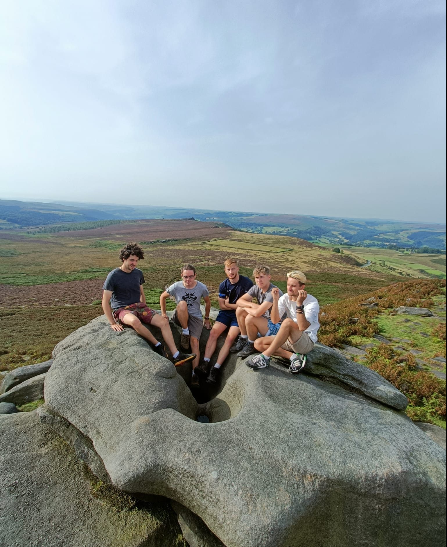 Male outdoor learning apprentices sat on a big rock with amazing scenic view of country side behind them - sun is shining