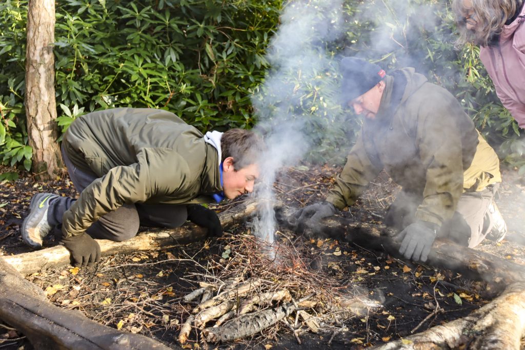 person leaning into fire in bushcraft activity blowing on smoke to light it more